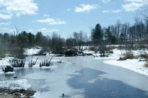 A serene winter landscape with a partially frozen pond, bare trees, and a clear blue sky with scattered clouds.