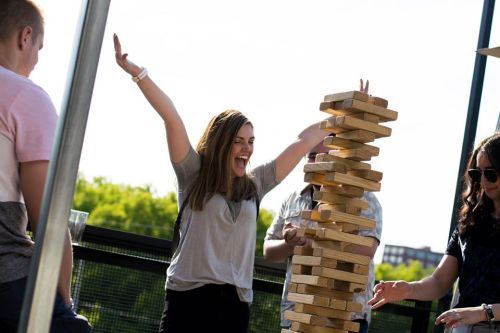 A woman celebrates with raised arms next to a tall Jenga tower, surrounded by friends outdoors.