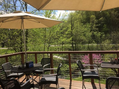 A sunny outdoor patio with tables and umbrellas, surrounded by lush greenery and a view of a winding road.