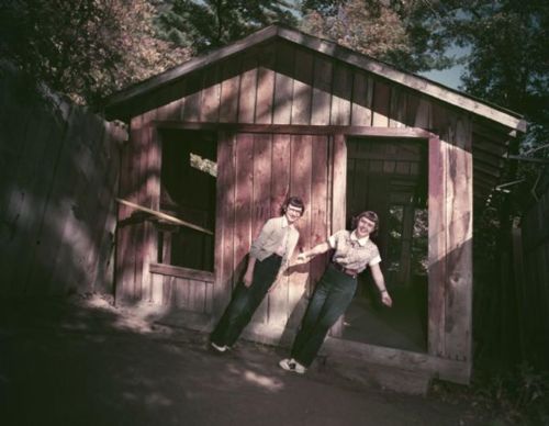 Two girls in vintage clothing stand playfully outside a wooden shed, surrounded by trees.