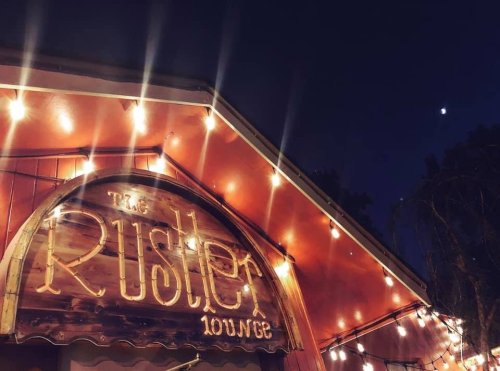 Sign for "The Rustler Lounge" illuminated by string lights at night, with a crescent moon visible in the sky.