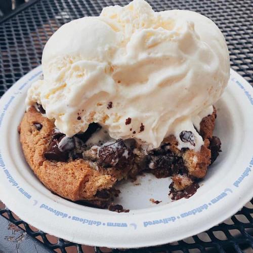A warm chocolate chip cookie topped with a generous scoop of vanilla ice cream on a paper plate.