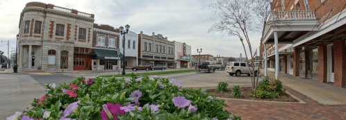 A street view of a small town with historic buildings, parked cars, and colorful flowers in the foreground.