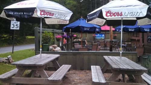 Outdoor seating area with picnic tables and umbrellas, surrounded by greenery and flowers, near a road sign.