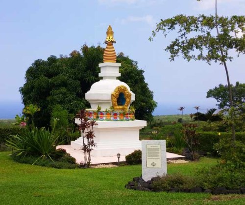 A white stupa with colorful details surrounded by lush greenery and a plaque in a serene outdoor setting.
