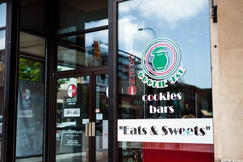A storefront window displaying "Cookie Jar" with signs for cookies, bars, and "Eats & Sweets." Open sign visible.
