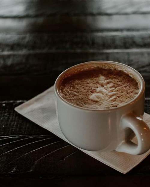 A white coffee cup on a napkin, filled with frothy coffee, resting on a dark wooden table.