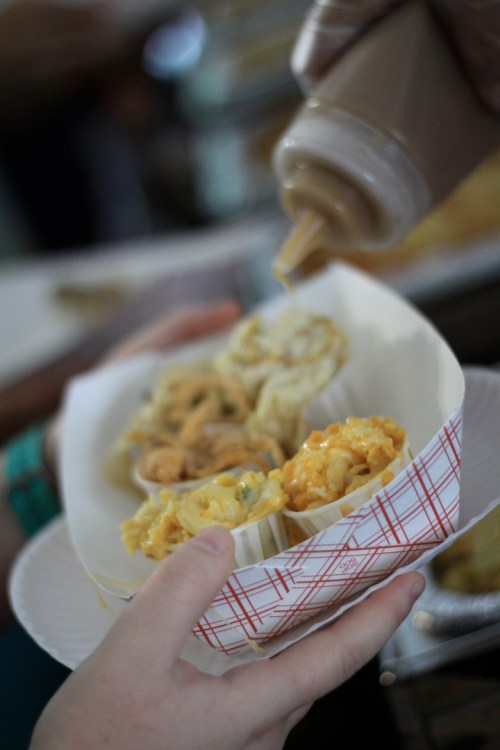 A hand holds a paper tray with three servings of macaroni and cheese, while a bottle of sauce is being poured over them.