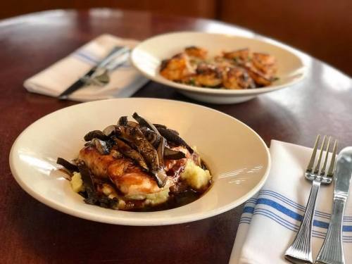 A plate of chicken atop mashed potatoes, garnished with green beans, served on a wooden table with another dish in the background.