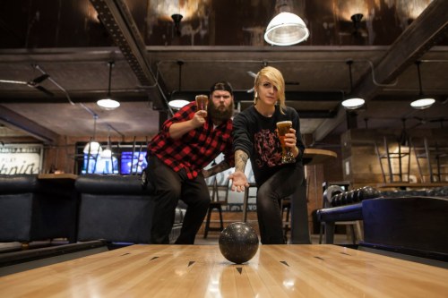 A man in a plaid shirt and a woman in a black sweater bowl together in a stylish, dimly lit bowling alley.