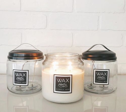 Three candle jars with black lids, one lit, labeled "WAX poetic CANDLE BAR," against a white brick background.