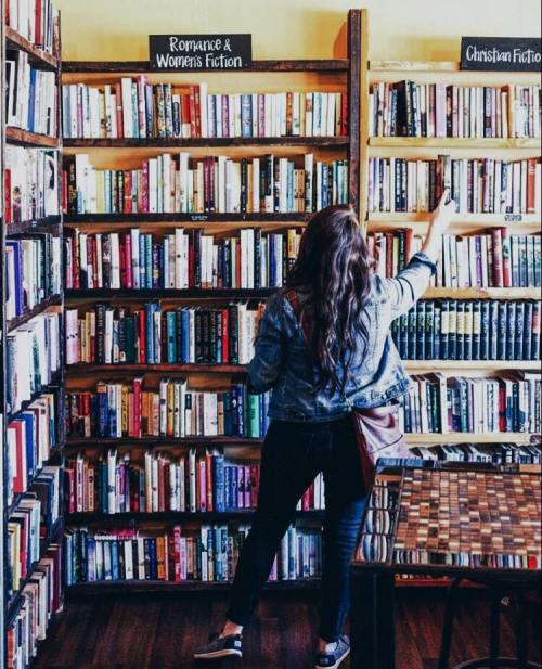 A person browsing books on shelves labeled "Romance & Women's Fiction" and "Christian Fiction" in a cozy bookstore.