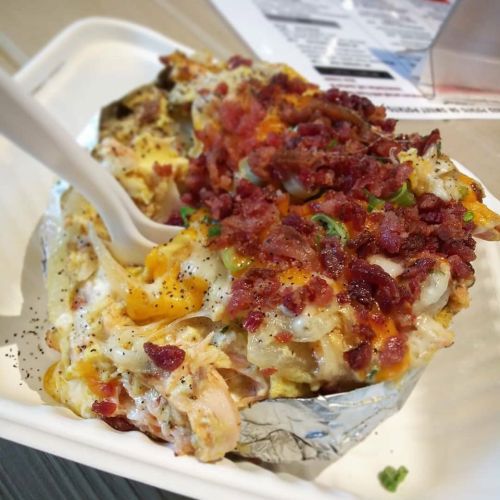 A loaded baked potato topped with cheese, bacon bits, and green onions, served in a white takeout container.