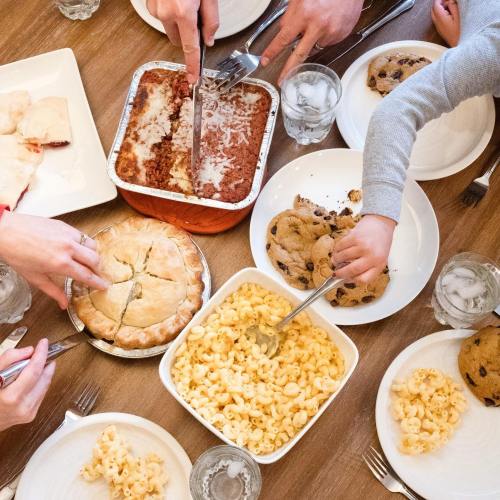 A table filled with various dishes: lasagna, macaroni, cookies, and pie, with hands serving food and drinks.