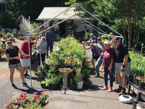 A group of people smiles and poses in a garden center, surrounded by plants and flowers under a metal frame.