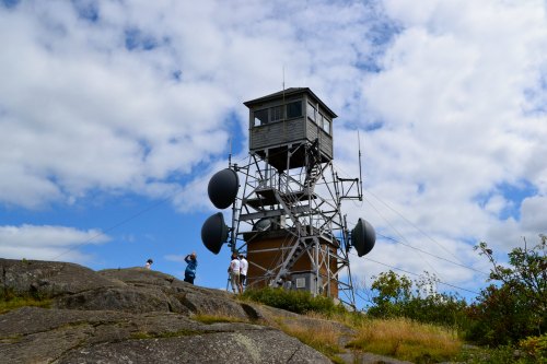 A tall communication tower on a rocky hill, with people nearby and a blue sky filled with clouds.