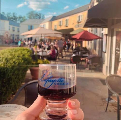 A hand holds a glass of red wine in front of a lively outdoor dining area with umbrellas and people enjoying the day.