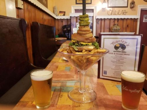 A towering burger with toppings sits in a glass, surrounded by fries, with two beers on a wooden table.