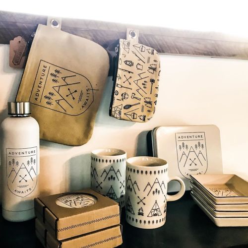 A collection of adventure-themed mugs, coasters, and pouches with outdoor designs displayed on a shelf.