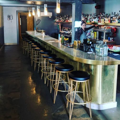 A sleek bar with a golden countertop, black stools, and a bartender arranging drinks in a stylish setting.