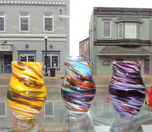 Three colorful glass vases with swirling patterns displayed in a window, with a street view in the background.