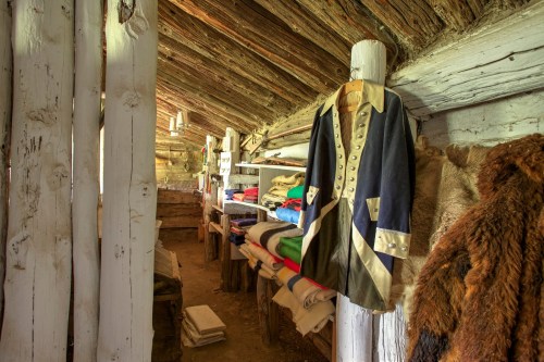Interior of a rustic cabin displaying historical clothing and neatly arranged blankets on wooden shelves.