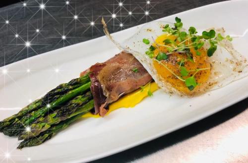 A gourmet dish featuring asparagus wrapped in ham, topped with a sunny-side-up egg and microgreens on a white plate.