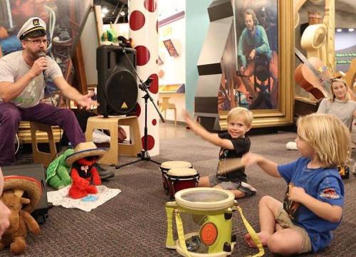 A man performs with children playing drums in a colorful, interactive space filled with toys and art.