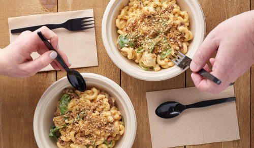 Two bowls of pasta with toppings, hands reaching in with forks and spoons, set on a wooden table with napkins.