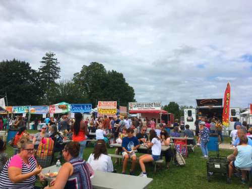 A bustling outdoor food festival with various food stalls and a large crowd enjoying the event under a cloudy sky.