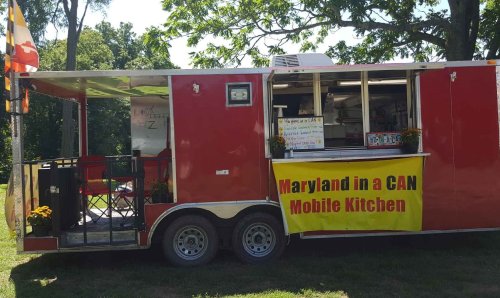 A red mobile kitchen trailer with a sign reading "Maryland in a CAN" and outdoor seating area.