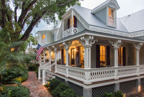 A charming house with a wraparound porch, surrounded by lush greenery and an American flag, illuminated at dusk.