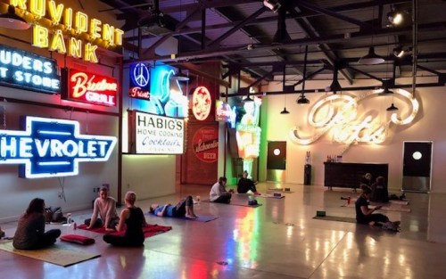 A yoga class in a spacious room adorned with colorful vintage neon signs. Participants are practicing on mats.