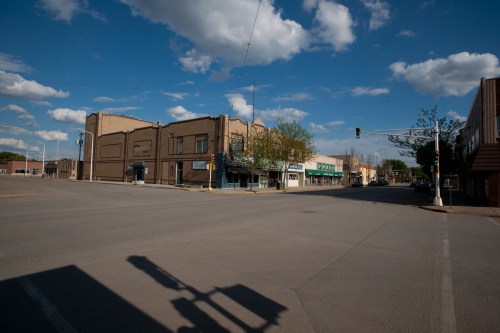 A quiet street scene featuring buildings, traffic lights, and a clear blue sky with scattered clouds.