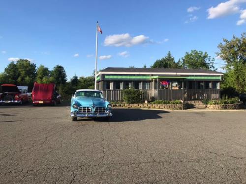 A vintage car parked in front of a diner with an "Open" sign, surrounded by classic cars and greenery under a blue sky.
