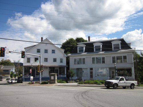 Two large buildings at an intersection, one with a porch, under a partly cloudy sky. A truck is parked nearby.
