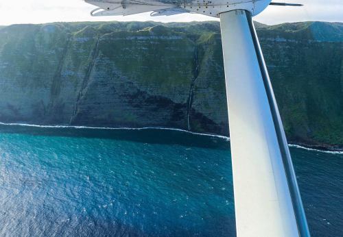Aerial view of a green cliffside meeting the ocean, with a plane wing in the foreground.