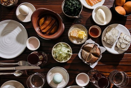 A wooden table set with various dishes, including eggs, greens, bread, and fruits, arranged for a meal.