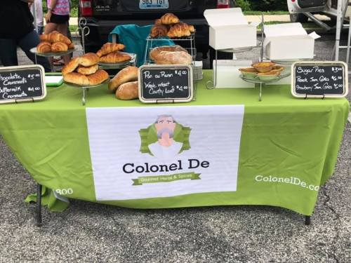 A market stall displaying various baked goods, including croissants and pastries, with a green tablecloth and signage for Colonel De.