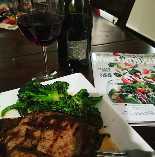 A plate with grilled steak and broccoli, a glass of red wine, and a magazine titled "Wine Industry" on a wooden table.