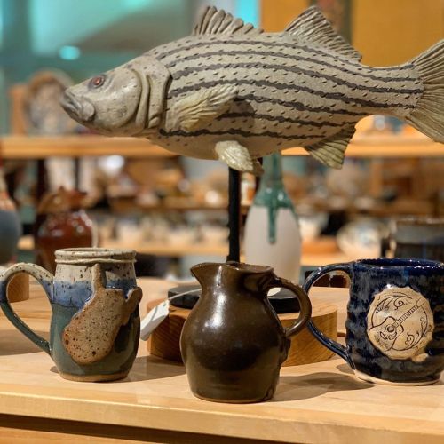 Three unique ceramic vessels and a decorative fish sculpture displayed on a wooden shelf in an art gallery.