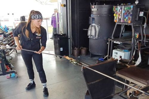A woman in a black outfit shapes molten glass using a long rod in a glassblowing studio.