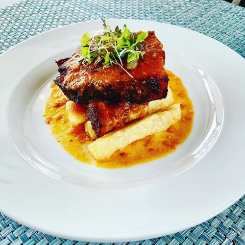 A plate of grilled meat atop mashed plantains, garnished with microgreens and served with a savory sauce.