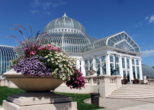A large glass conservatory with a dome, surrounded by colorful flower beds and a clear blue sky.