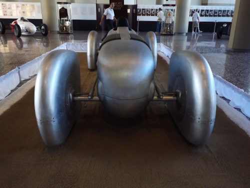 A vintage silver racing car displayed indoors, featuring large wheels and a sleek, aerodynamic design.