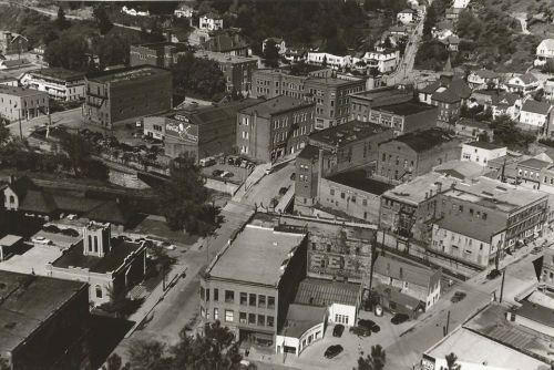 Aerial view of a historic town with buildings, streets, and parked cars, showcasing a mix of architecture and urban layout.