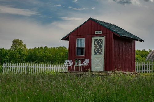 A small red cabin with a white door and chairs on a grassy field, surrounded by trees and a white picket fence.