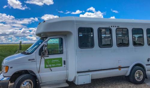 A white shuttle bus parked on a rural road under a blue sky with fluffy clouds. Green logo visible on the side.