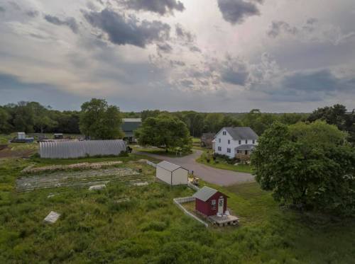 Aerial view of a farm with green fields, a red shed, and a white house under a cloudy sky.