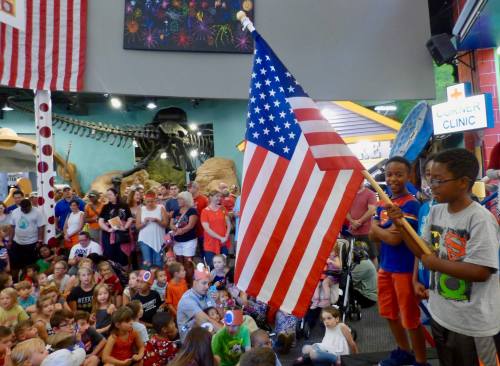 A group of children and adults gather to celebrate, with one boy holding an American flag on stage.